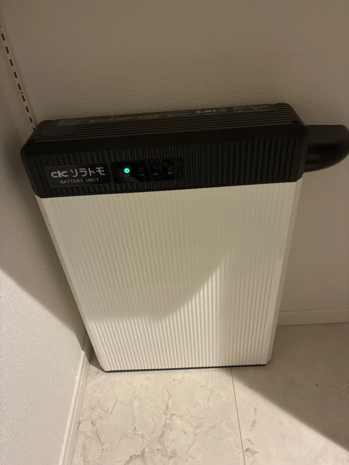 長州産業の蓄電池ユニット CB-LMK65A。脱衣所の床に置かれた白い本体と「ソラトモ BATTERY UNIT」の表示、運転ランプが緑に点灯