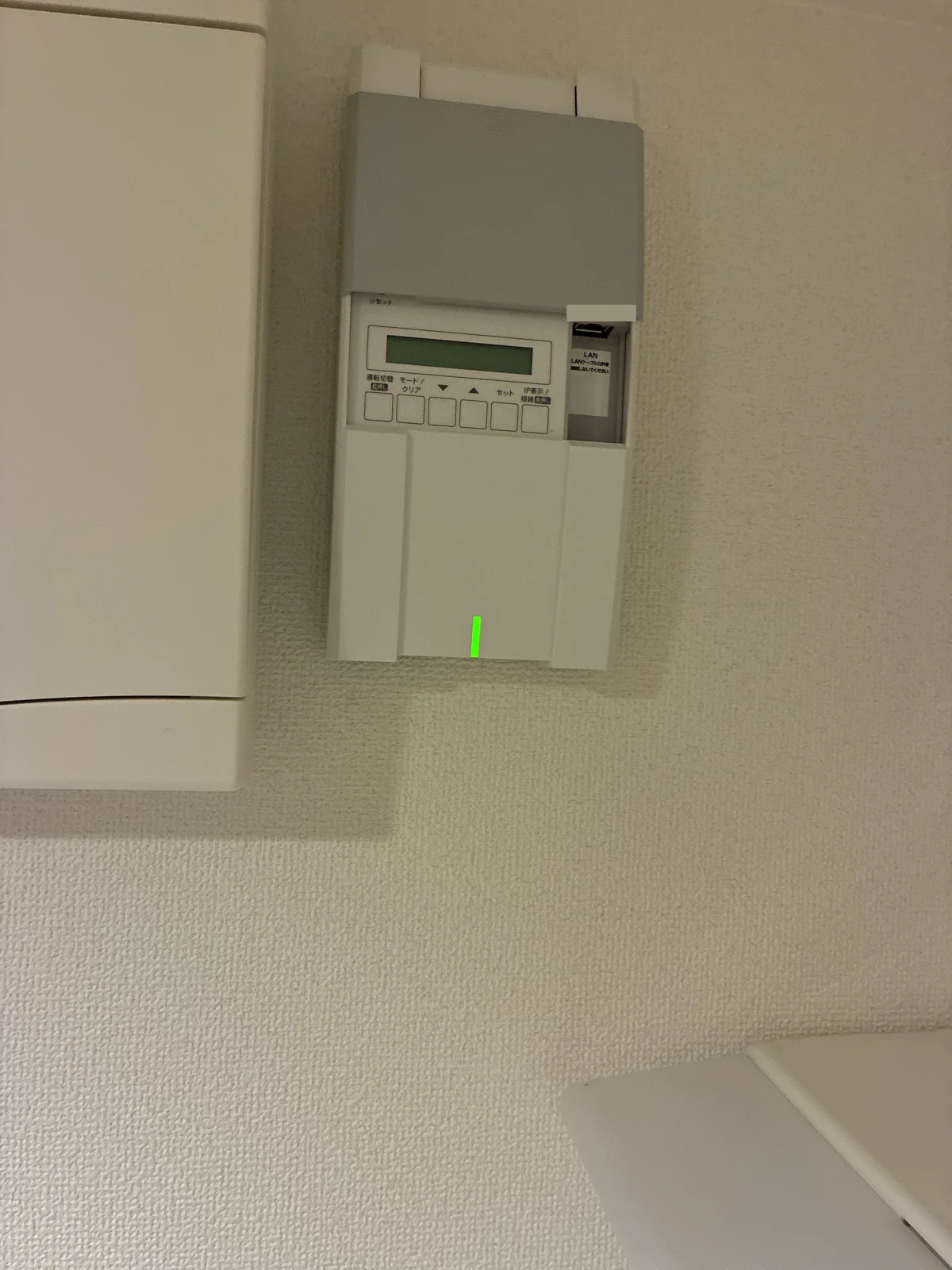 脱衣所の壁に付いている計測ユニット MHCS-M02A(右)と特定負荷用分電盤(左の白い箱)。計測ユニットの緑のランプが通常運転の合図