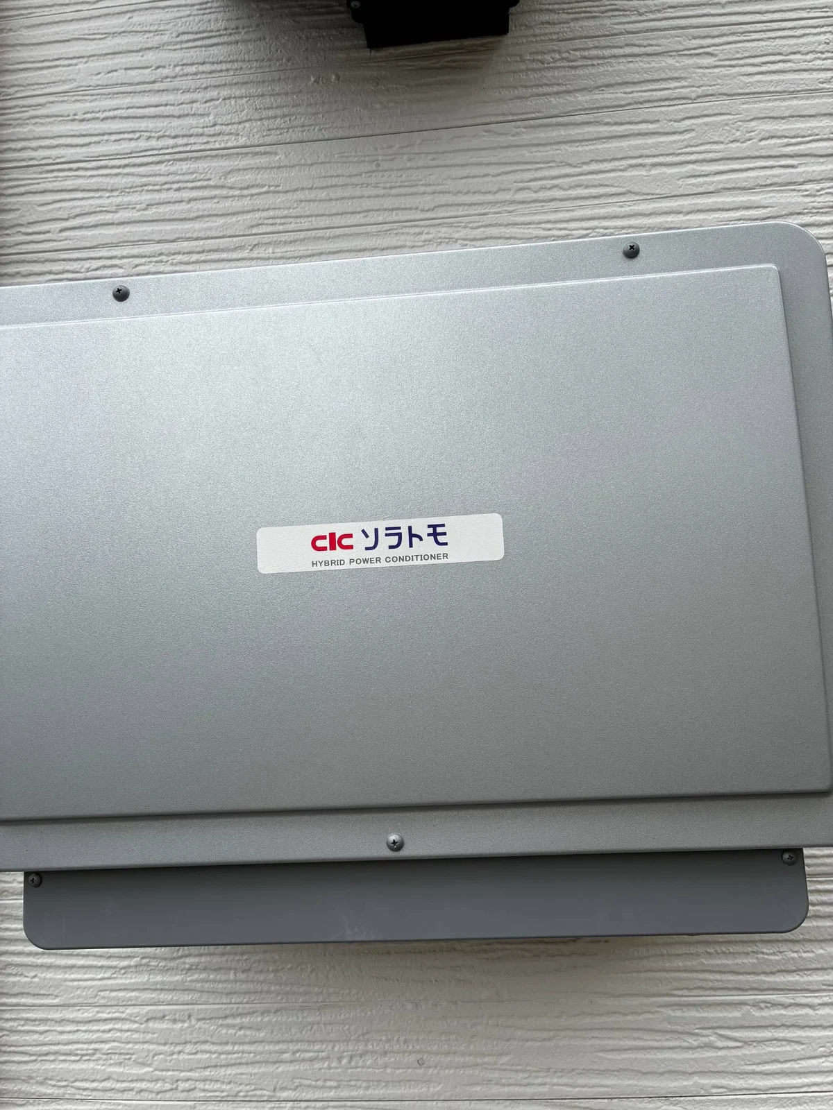 我が家のパワコン。外壁に付いた銀色の箱で「ソラトモ HYBRID POWER CONDITIONER」のシールが貼ってある